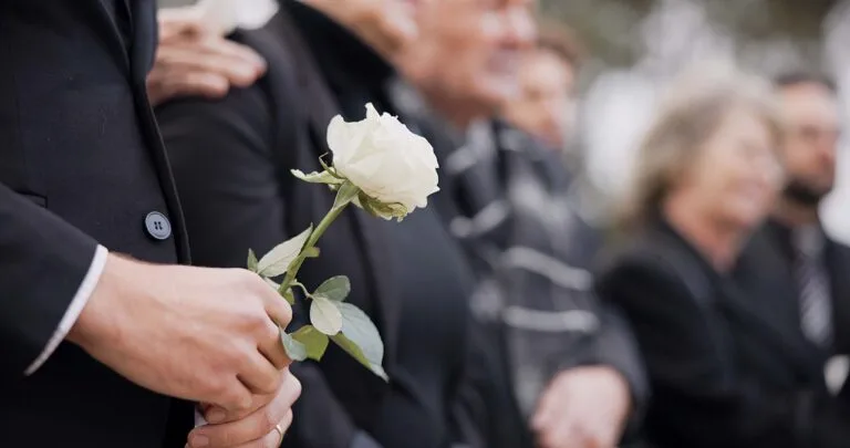 Hands, rose and a person at a funeral in a cemetery in grief while mourning loss at a memorial service. Death, flower and an adult in a suit at a graveyard in a crowd for an outdoor burial closeup